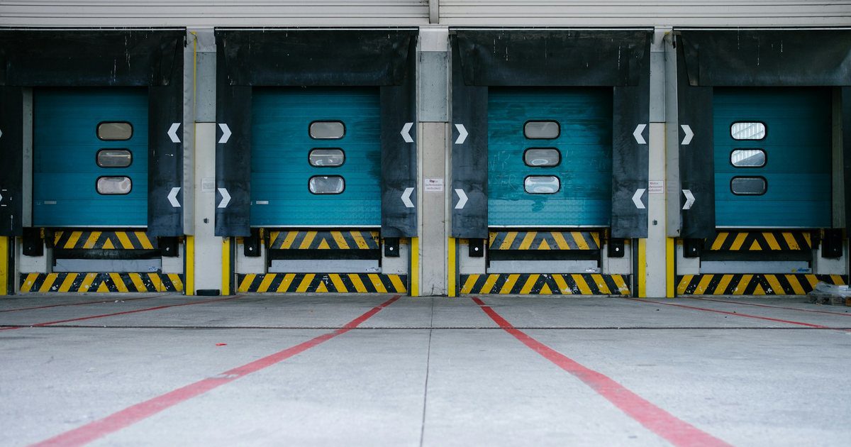 A long industrial wall of identical closed roller shutters receding into the distance, every one of them shut.