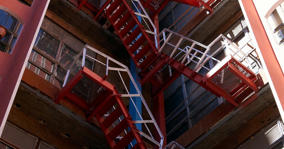 An exterior steel staircase climbing flight after flight up the flank of a large concrete industrial building.