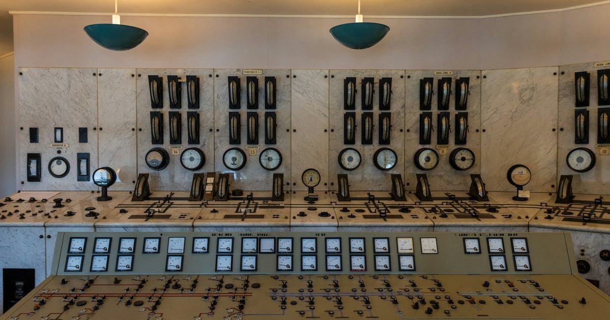 An empty industrial control room, banks of gauges and labelled switches lit and running with no operator at the desk.