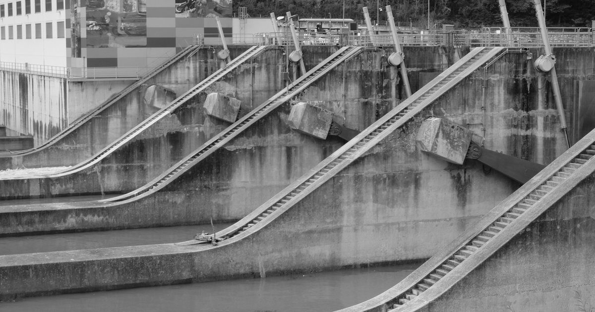 A concrete dam spillway with a row of identical closed gates, one of them open and discharging water.