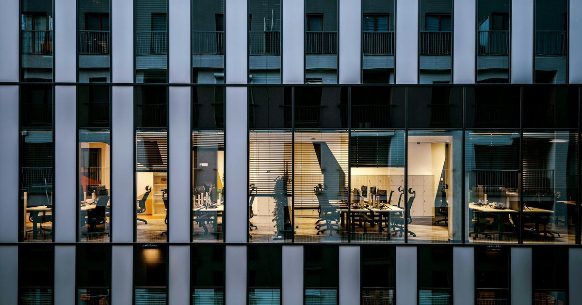 A tall building facade at dusk, hundreds of identical windows, a scattered few lit and most of them dark.